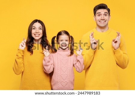Young happy parents mom dad with child kid girl 7-8 years old wear pink knitted sweater casual clothes wait special moment keep fingers crossed isolated on plain yellow background. Family day concept.