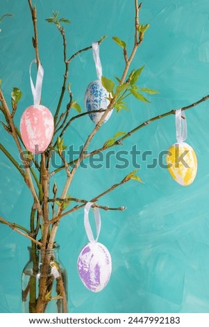 Holiday Easter image of spring twigs with young leaves in simple glass vase decorative pastel color eggs hanging on them on turquoise background. Creative Easter card. Vertical format. Closeup. Royalty-Free Stock Photo #2447992183
