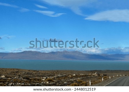 Panoramic photography of a lake and a snowy mountain range behind with a cloudy clear sky beside a road