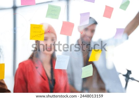 Business team uses a glass wall covered with sticky notes to outline processes during meeting. Engaged colleagues gather around a glass brainstorming board. plotting ideas and strategies. Royalty-Free Stock Photo #2448013159