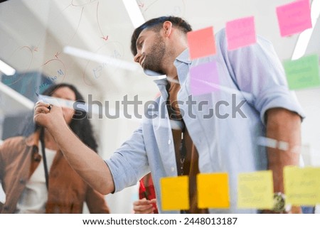 Business team uses a glass wall covered with sticky notes to outline processes during meeting. Engaged colleagues gather around a glass brainstorming board. plotting ideas and strategies. Royalty-Free Stock Photo #2448013187