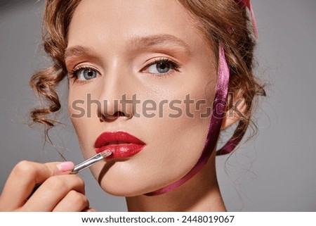 A young woman in a studio setting on a grey background, applying lipstick to enhance her lips with precision and care.