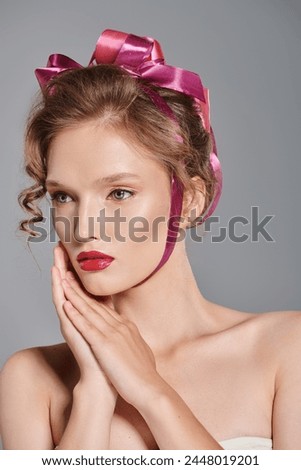 A young woman with a pink bow in her hair poses gracefully in a studio, exuding classic beauty on a grey background.