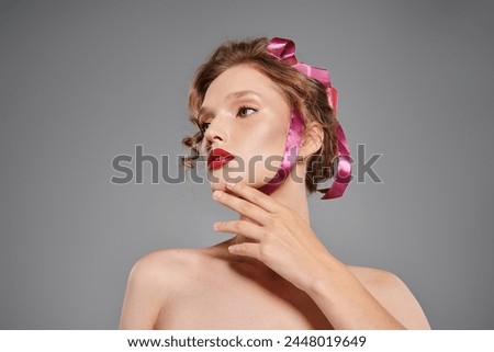 Young woman exudes classic beauty as she confidently poses with a pink bow on her head in a studio setting on a grey background.