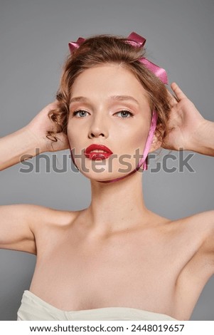 A young woman with classic beauty strikes a pose in a studio, wearing a pink bow on her head against a grey background.
