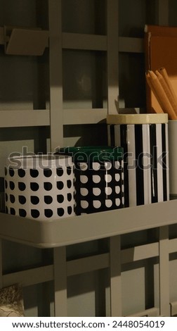 A closeup of the storage shelves, featuring a white metal shelf with black and green plastic cans of different sizes on it. The surfaces have grid designs with polka dots and stripes in various shades