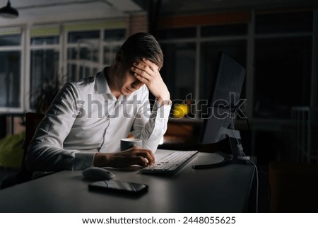Tired male office worker working late at night, using desktop computer and touching forehead. Exhausted businessman working on laptop computer until night. Concept of stressful life and deadline. Royalty-Free Stock Photo #2448055625