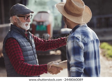 Two farmers young and mature shaking hands in front of tractor on cow farm as sign of deal.