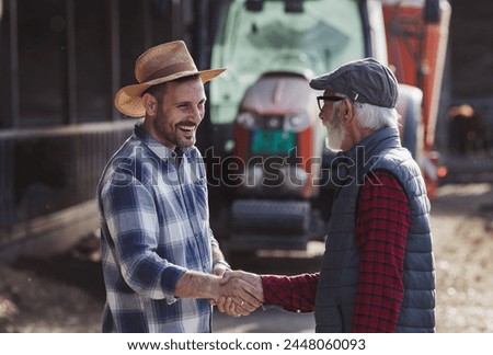 Two farmers young and mature shaking hands in front of tractor on cow farm as sign of deal.