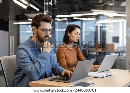 Two focused professionals working together at a desk with laptops in a contemporary office setting, conveying collaboration and teamwork.