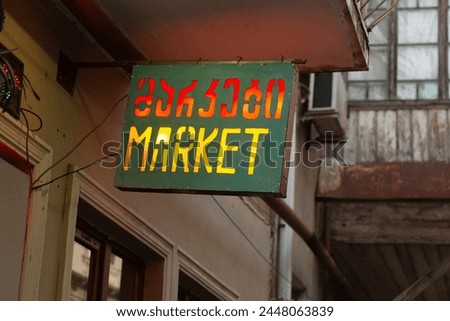 Street photo. Horizontal green sign for grocery store, supermarket, shop with glowing red and yellow letters on facade of house. Tbilisi city. Sale, trade, pay. Text. Translation from Georgian: Market