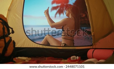 Female resting on campsite relaxing near ocean. Young woman sitting outside the tent at sunset on the beach taking selfie pictures on smartphone, vacation outdoors.
