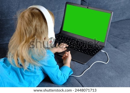 green screen mockup, child 2-3 years old, girl in headphones lies on sofa front laptop, uses Internet, watches educational cartoons, plays computer games, Parenting in digital age, remote learning