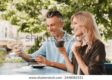 Happy caucasian middle-aged spouses couple hugging embracing bonding and drinking coffee in cafe restaurant while watching social media on cellphone together on a date. Love and relationship concept.
