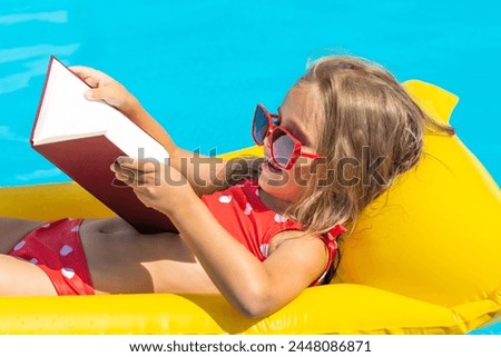 Cute little girl reading children book on inflatable mattress in swimming pool with blue water on warm summer day on tropical vacations. Summertime relax. Cute little girl sunbathing on air mattress