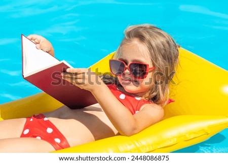 Cute little girl reading children book on inflatable mattress in swimming pool with blue water on warm summer day on tropical vacations. Summertime relax. Cute little girl sunbathing on air mattress