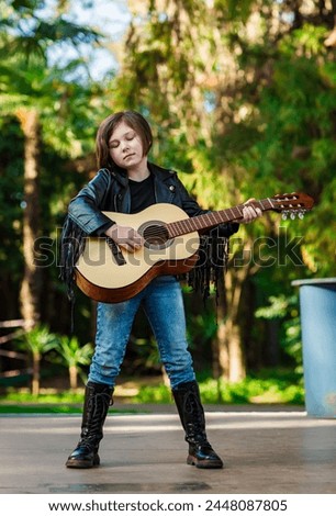 A beautiful little girl, in rock style,in a leather jacket and jeans,stands on a summer stage, and plays an acoustic guitar
