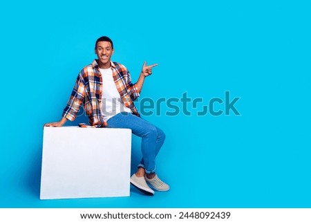 Full size photo of young man sit cube direct fingers empty space wear shirt isolated on blue color background