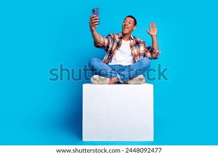 Full size photo of nice young man sit podium phone make selfie arm wave hi empty space wear shirt isolated on blue color background