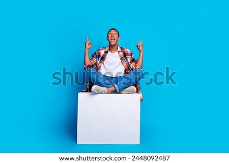 Full size photo of young man sit cube indicate fingers up empty space wear shirt isolated on blue color background