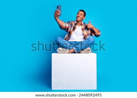 Full size photo of nice young guy make selfie show thumb up sit podium empty space wear shirt isolated on blue color background