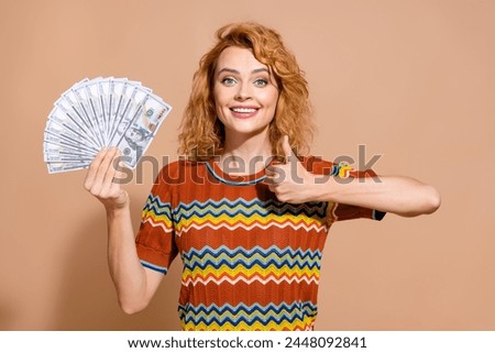 Photo portrait of lovely young lady hold money dollars show thumb up dressed stylish striped garment isolated on beige color background