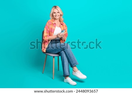 Portrait of cheerful nice retired woman wear plaid trendy clothes sit chair hold iphone isolated on cyan color background