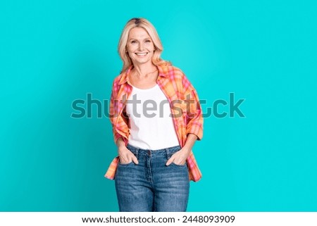 Photo of toothy beaming retired woman straight hair dressed checkered shirt holding hands in pockets isolated on teal color background
