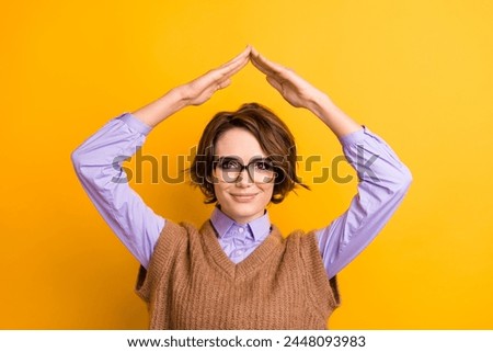 Photo of satisfied girl dressed knitwear waistcoat in glasses holding hands like roof over head isolated on yellow color background