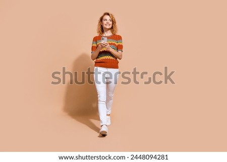 Full size photo of optimistic girl wear ornament t-shirt walk with smartphone look at offer empty space isolated on beige color background