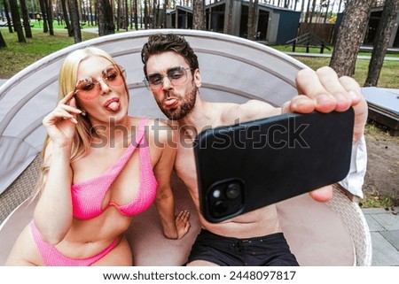 Happy young man and woman grimacing while making selfie on the mobile phone, sitting on the sunbed. Summer vacation, relationship, lifestyle concept