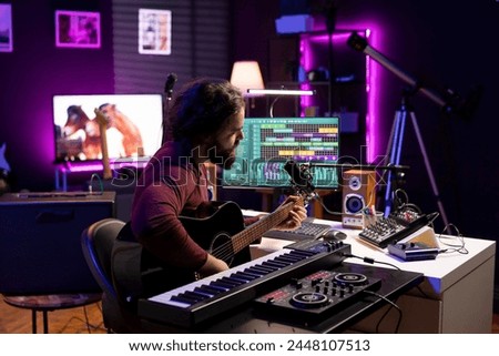 Musical artist recording his acoustic guitar accords in home studio, creating a beautiful song by singing lyrics on microphone. Producer uses mixing console and daw software to compose tunes.