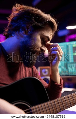 Stressed sleepy artist practicing acoustic instrument in his home studio, recording new tunes and editing it in post production. Young artist playing guitar with frustration, deals with burnout.