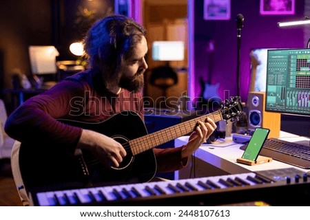 Musician playing acoustic instrument next to greenscreen on smartphone, learning new accords on strings to play on guitar. Artist producer developing his musical skills in his home studio.