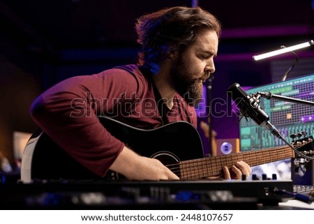 Songwriter composing a new song on acoustic guitar in home studio, using a microphone to sing the chords. Artist works with mixing console and daw software, guitarist producing new music.