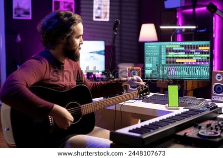 Musician learning to tune his guitar with internet tutorial via greenscreen on phone, twisting the knobs and practicing music lessons. Artist songwriter does rehearsal with acoustic instrument.