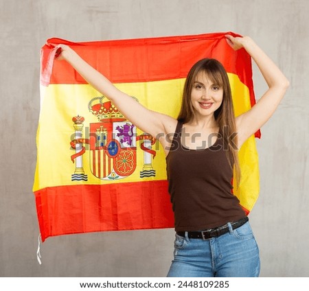 Happy young woman with large flag of Spain posing gladly against light unicoloured background