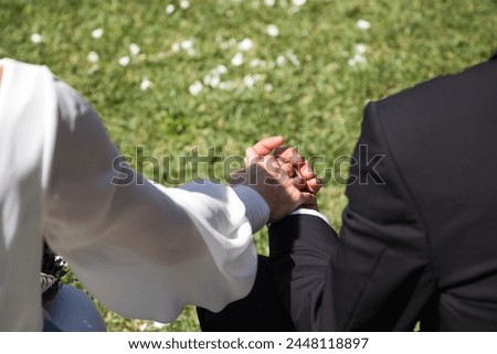 Detail of clasped hands of newly married couple dressed as bride and groom, he in black suit and she in white suit. Photo taken from behind Royalty-Free Stock Photo #2448118897
