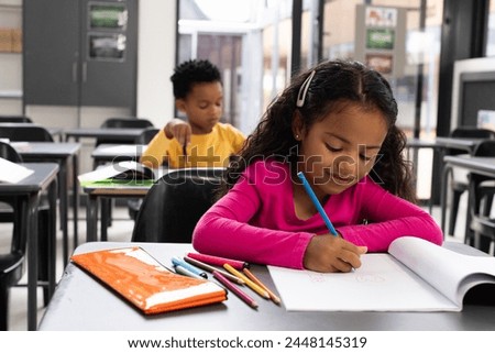 Biracial girl with curly brown hair is in a school classroom, African American boy in the background. They are focused on their tasks in a brightly lit classroom setting. Royalty-Free Stock Photo #2448145319