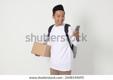 Portrait of happy Asian muslim man holding mobile phone while carrying cardboard box with excited expression. Going home for Eid Mubarak. Isolated image on white background