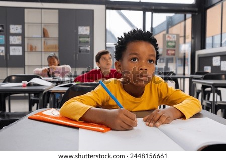 African American boy focused on his schoolwork in a school classroom setting. Biracial girl and another child are in the background, engaged in their studies.