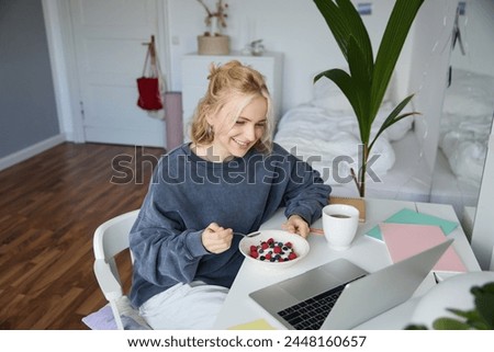 Image of smiling young woman watching videos on laptop while eating breakfast and drinking tea, looking at screen, sitting in her room.