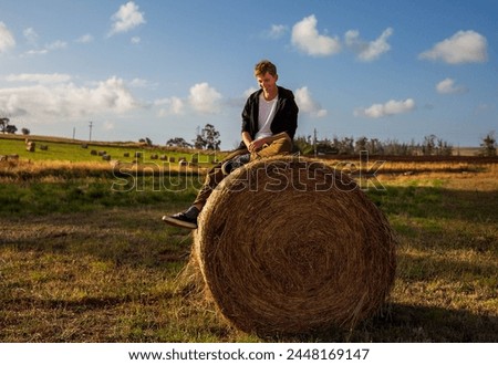Portrait of handsome young guy sitting on top of huge haystack on farm.