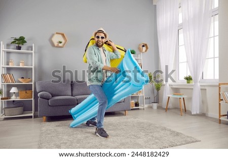 Funny overjoyed excited man holding inflatable mattress and suitcase standing in living room at home and having fun. Happy tourist is going on summer holiday trip. Vacation concept.