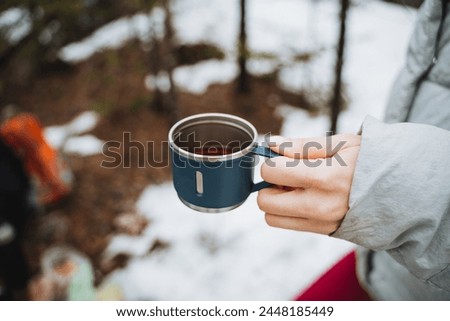 Woman holding a glass of tea on the background of snow, drinking tea in a wild forest, trekking in nature, traveling in spring in cold weather, a glass of coffee. High quality photo