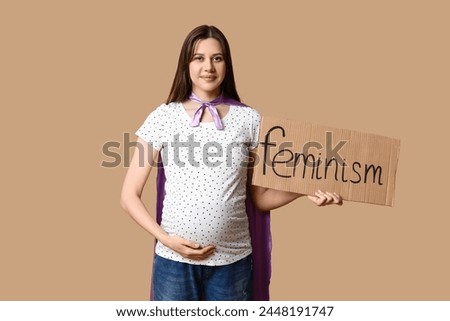 Beautiful young pregnant woman in superhero cape holding cardboard with word FEMINISM on brown background