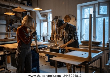 Carpentry workshop. Focused spouses woman man working in carpentry workshop assembling table, joining base and legs. Working together on an individual order. Furniture manufacturing, small business Royalty-Free Stock Photo #2448224813