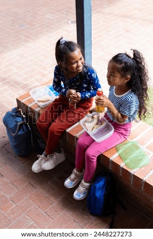 Two biracial girls enjoy a lunch break outdoors at school, sharing smiles and sandwiches. Dressed in casual attire, they sit on a brick ledge with lunchboxes and a blue backpack nearby. Royalty-Free Stock Photo #2448227273