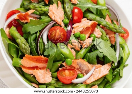 smoked salmon salad with asparagus, rocket salad, green peas, tomatoes and red onion in white bowl on white wooden table, close-up, dutch angle view Royalty-Free Stock Photo #2448253073