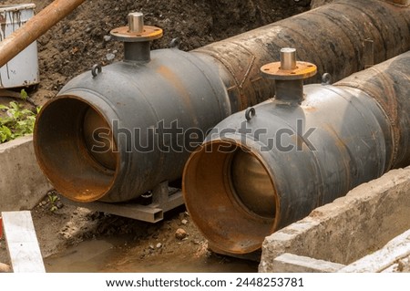 Two black pipes with a rusty top and a metal ring. The pipes are next to each other and are in a muddy area
